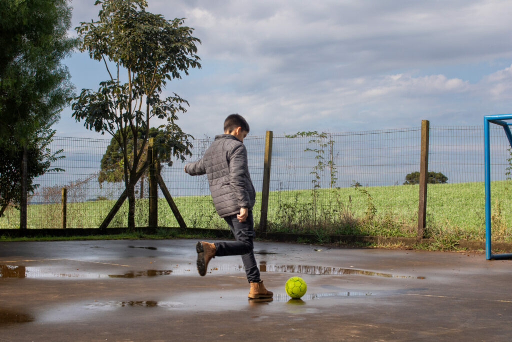 Un niño juega al fútbol en la escuela rural Attilio Benedetti de Vacaria, rodeada de plantaciones de soja. Un cultivo de cobertura que fertiliza el suelo, preparándolo para la siembra de soja, se puede ver entrando al patio de la escuela. Créditos Anna Ortega