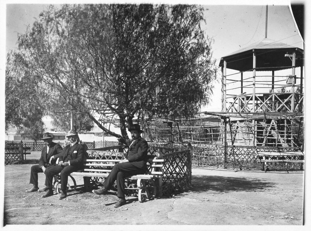 Retrato de grupo de hombres sentados en una banca, bajo un árbol, en la plaza de la oficina salitrera. Créditos: Colección Fotografía Museo Histórico Nacional.