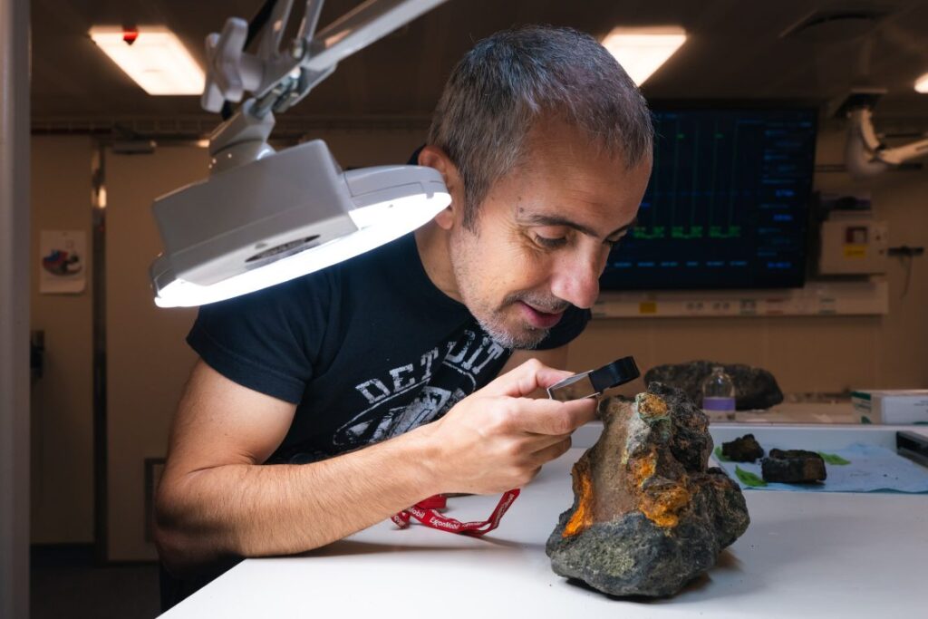 El científico jefe Aaron Micallef, del Monterey Bay Aquarium Research Institute, examina una roca procedente de un depósito de sulfuros masivos. Aunque las fuentes hidrotermales son estructuras individuales con una vida relativamente breve, de apenas unos miles de años, estos depósitos de sulfuros masivos se forman a una escala mucho mayor y durante períodos considerablemente más prolongados. Son estructuras de gran relevancia, poco frecuentes y de especial interés para el equipo científico. Créditos: Alex Ingle, Schmidt Ocean Institute.